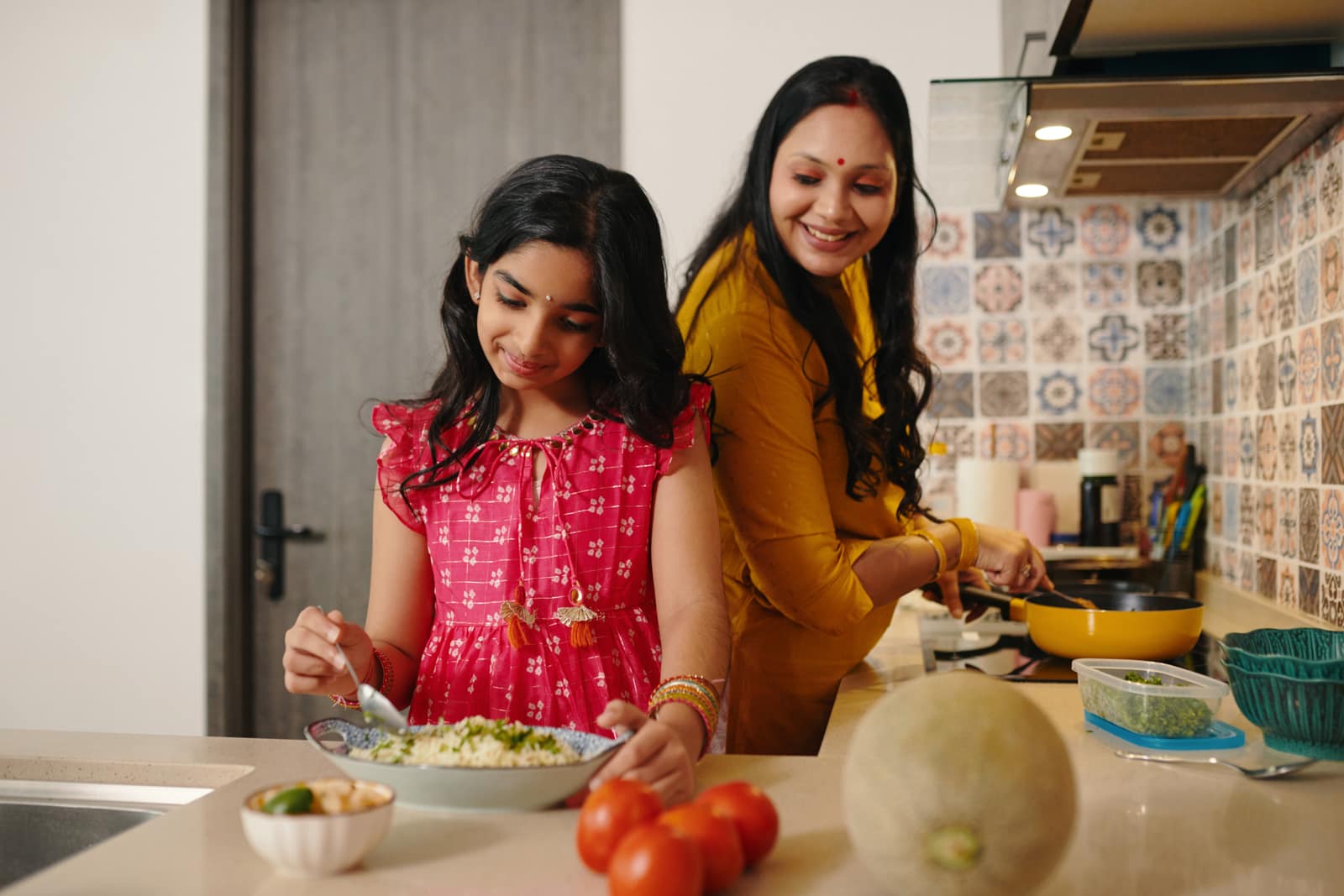 A mother and her daughter preparing a meal together at a kitchen counter, fresh vegetables in front of them and a pan on the hob behind