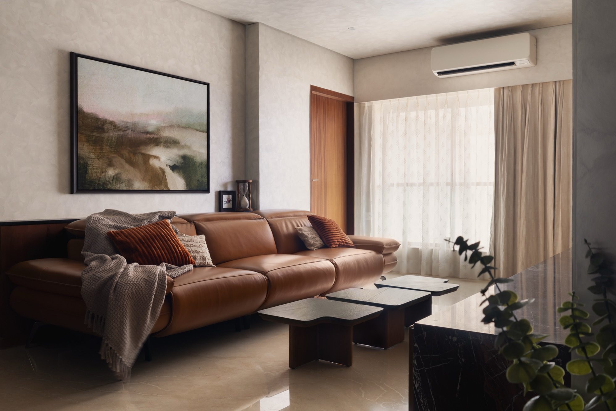 Plum House living room — a cognac leather recliner, sculptural nesting coffee tables and textured off-white plaster walls, daylight through sheer curtains