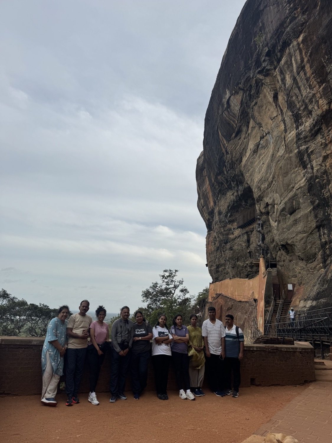 The group standing on the red-earth terrace beneath Sigiriya's dramatically overhanging western rock face, with the metal stairway to the summit clinging to the cliff at the right