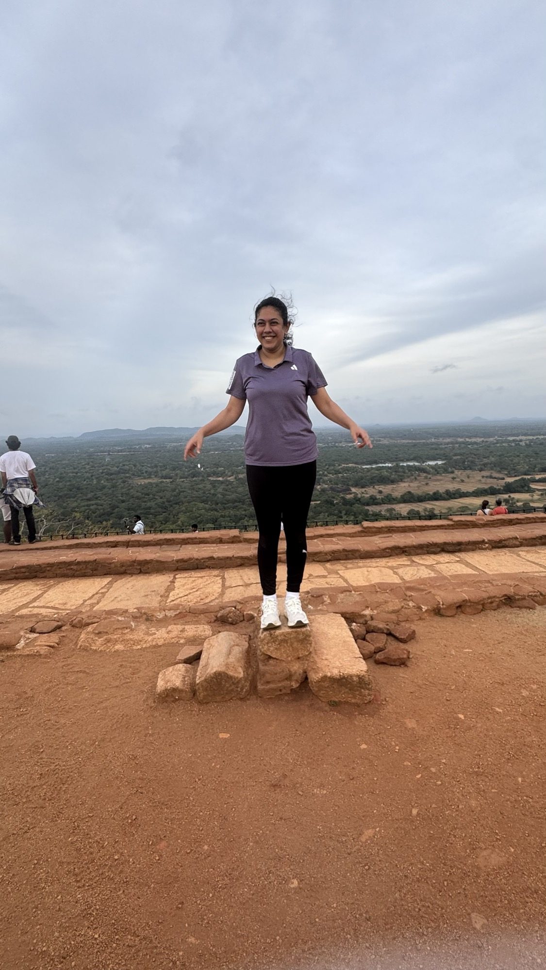 Digital Editor Inchara R Y standing on the summit of Sigiriya, arms open, with the vast green dry-zone plain and a distant reservoir spread out far below