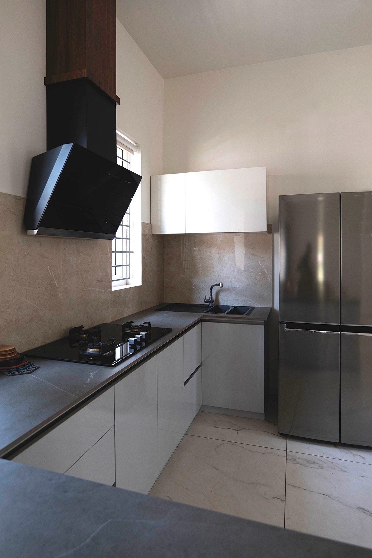 The kitchen — a hob beneath a black chimney hood, a window at the worktop, white cabinets, a sink at the return and a steel refrigerator