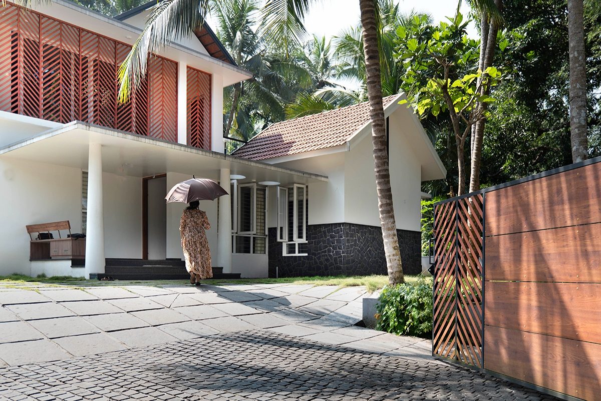 The approach to The Courtyard — a patterned timber screen on the upper level, a sheltered entrance with white columns, coconut palms and a woman with an umbrella