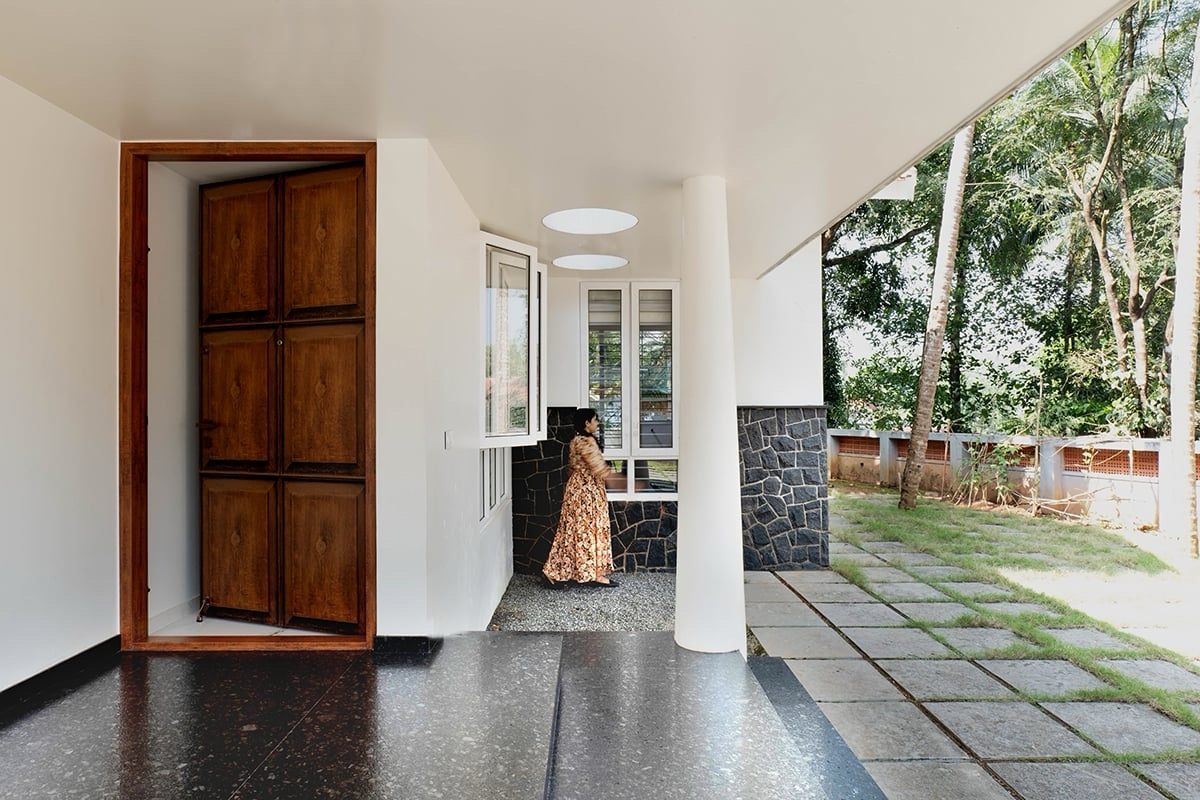 The entrance sit-out — a timber door, a white column and two circular openings in the overhead slab, with a dark floor and the garden beyond