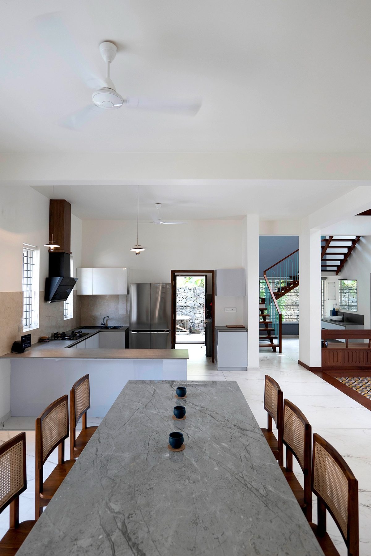 View from the dining table — a long stone-topped table with chairs, the open kitchen, pendant lights, a rear doorway and the staircase