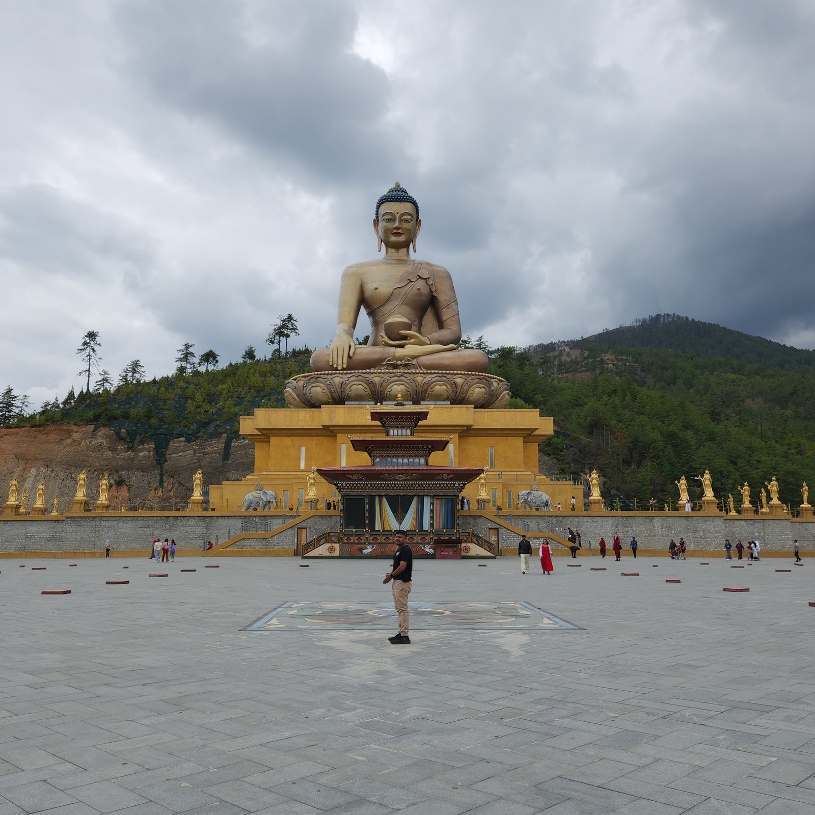 Amogh N P standing alone in the middle of the great paved court of the Buddha Dordenma, the gilded statue seated above its painted throne hall, with gilded figures along the court and a forested ridge under heavy cloud