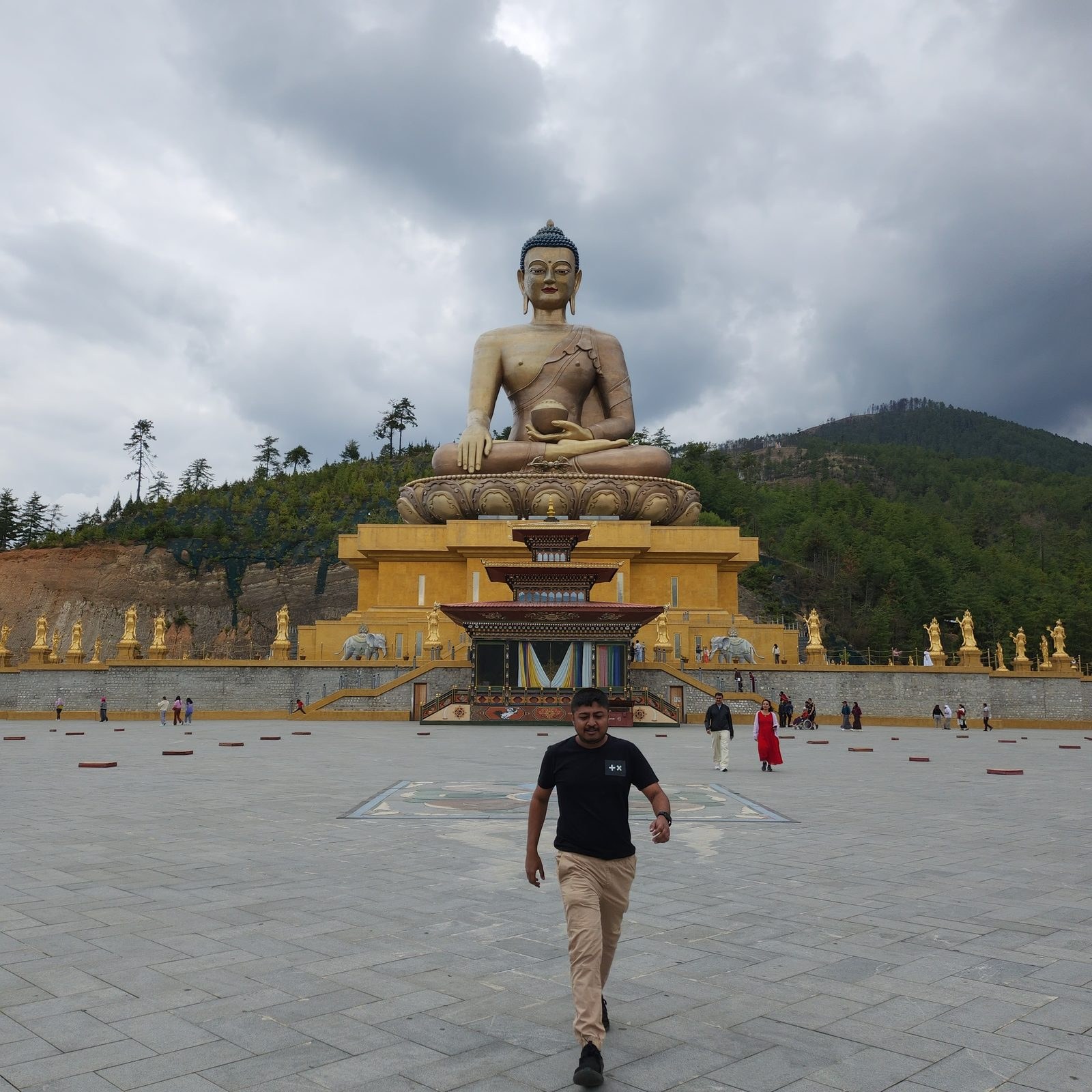 Amogh N P walking across the paved court toward the Buddha Dordenma, the statue seated above its throne hall against a forested ridge and heavy cloud