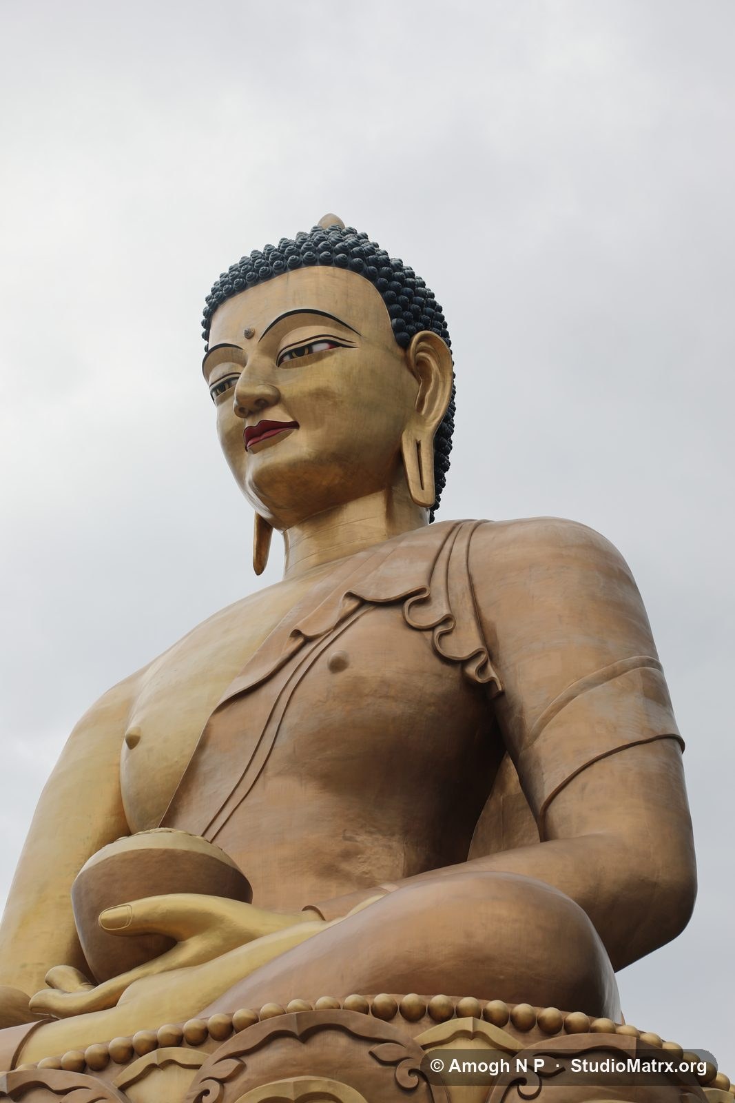 The bronze face and shoulders of the Buddha Dordenma against a pale clouded sky