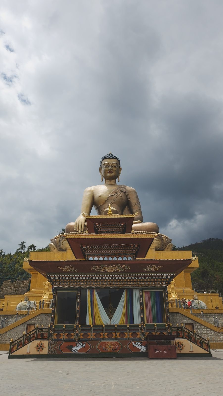 The Buddha Dordenma seated above its painted throne hall, seen straight on from the court under dark cloud