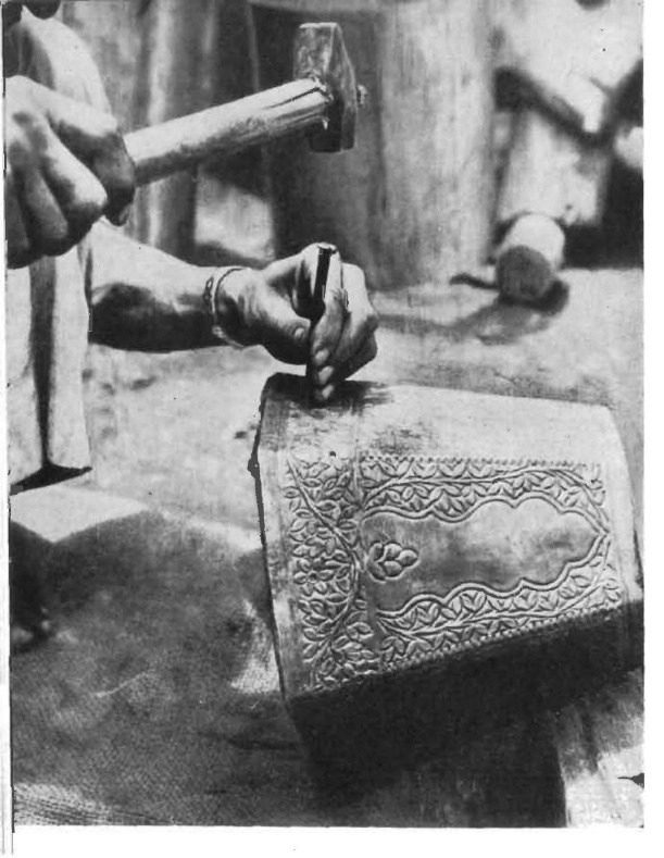 Black-and-white archival photograph: a craftsperson's hands holding a chisel and hammer, chasing a pattern into a brass vessel