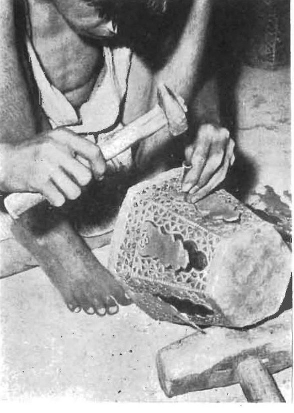 Black-and-white archival photograph: a seated craftsperson striking a tool with a hammer to pierce a pattern into a metal object