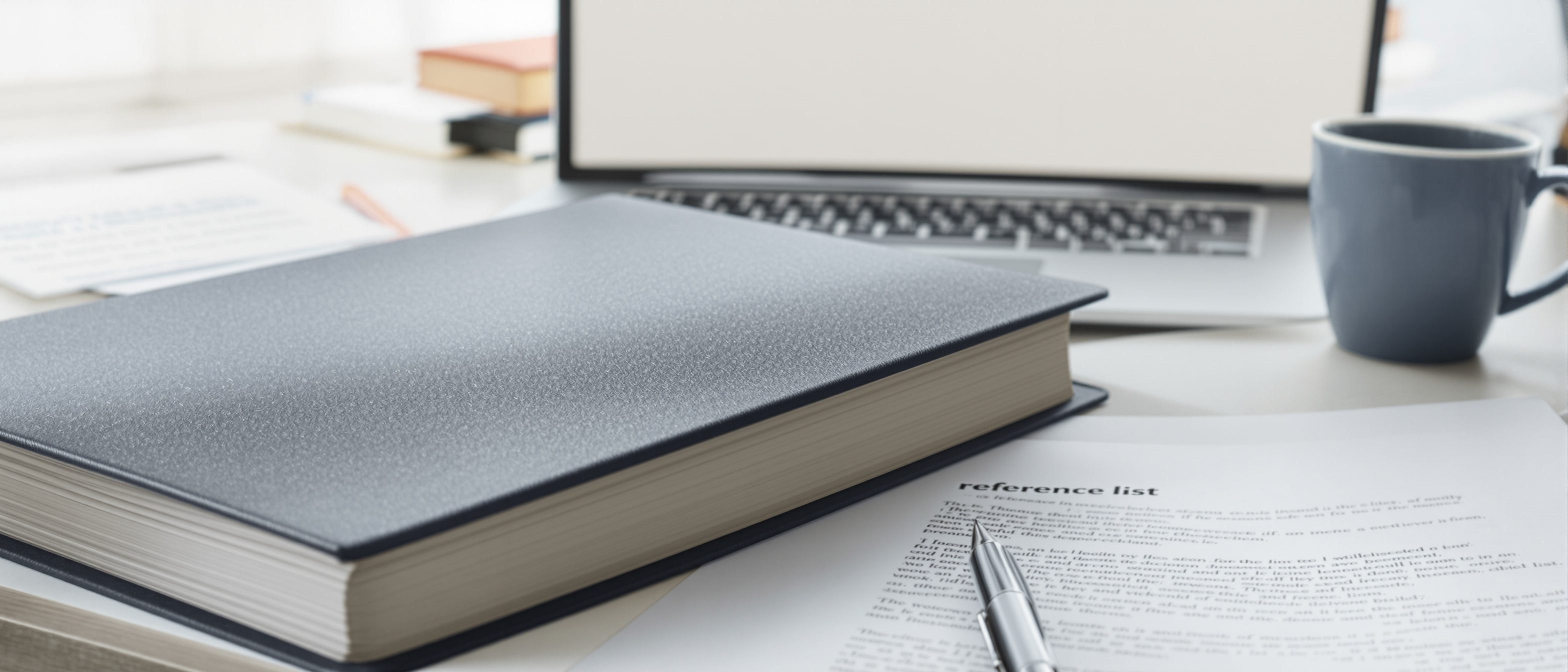 A bound architecture dissertation and a laptop open to a draft on a tidy desk, with a printed reference list and a coffee cup, the writing-up stage of research.