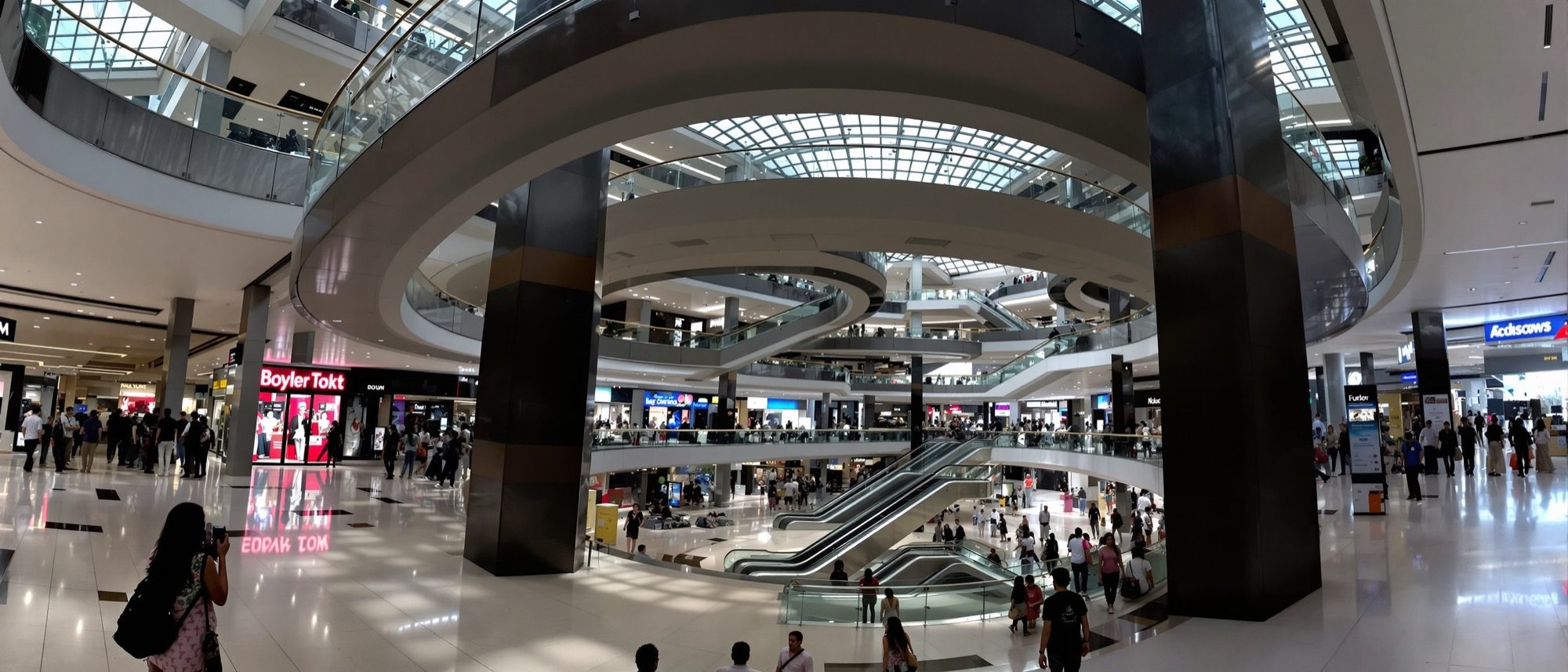 The daylit multi-level atrium of a modern shopping mall — shops linked by escalators under a glazed skylight, the magnet-and-generator type at work.