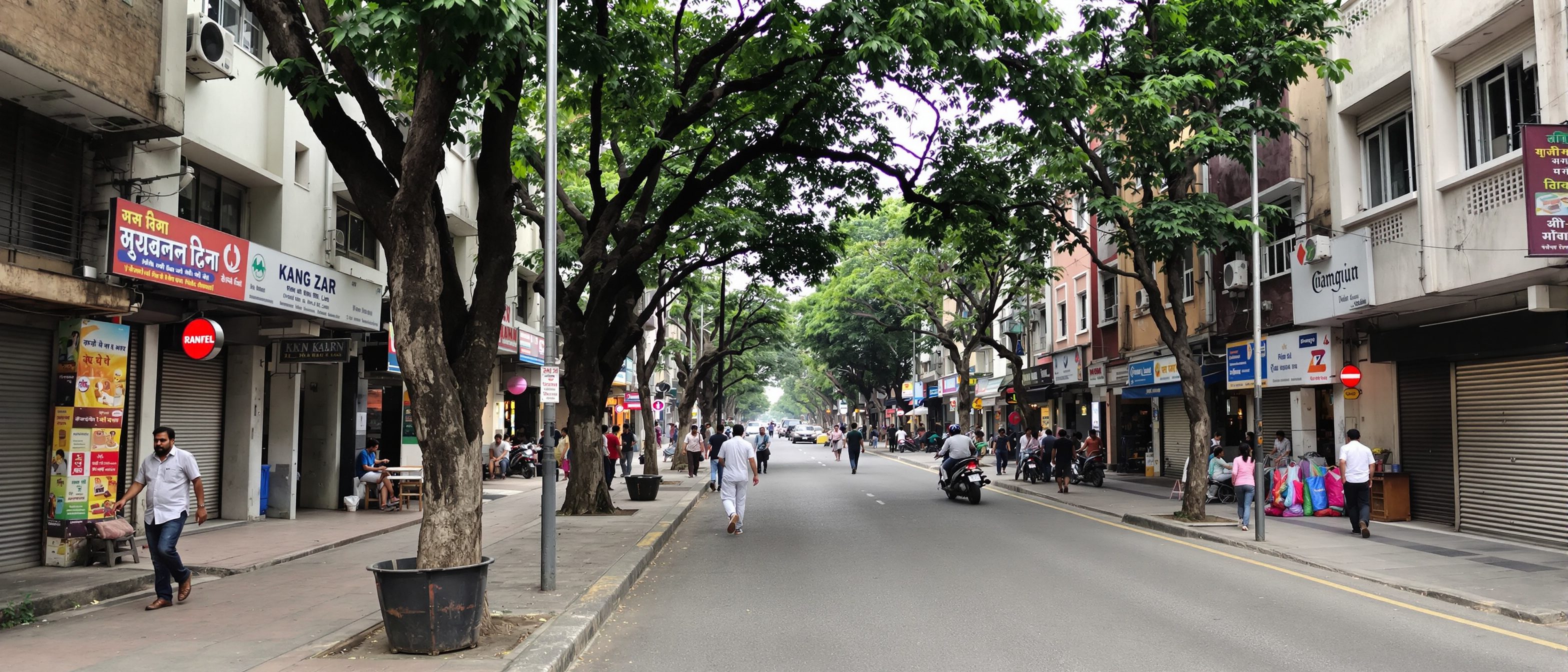 A well-enclosed mixed-use urban street in an Indian city with continuous building frontages, active ground-floor shops, street trees and people walking on a wide footpath.