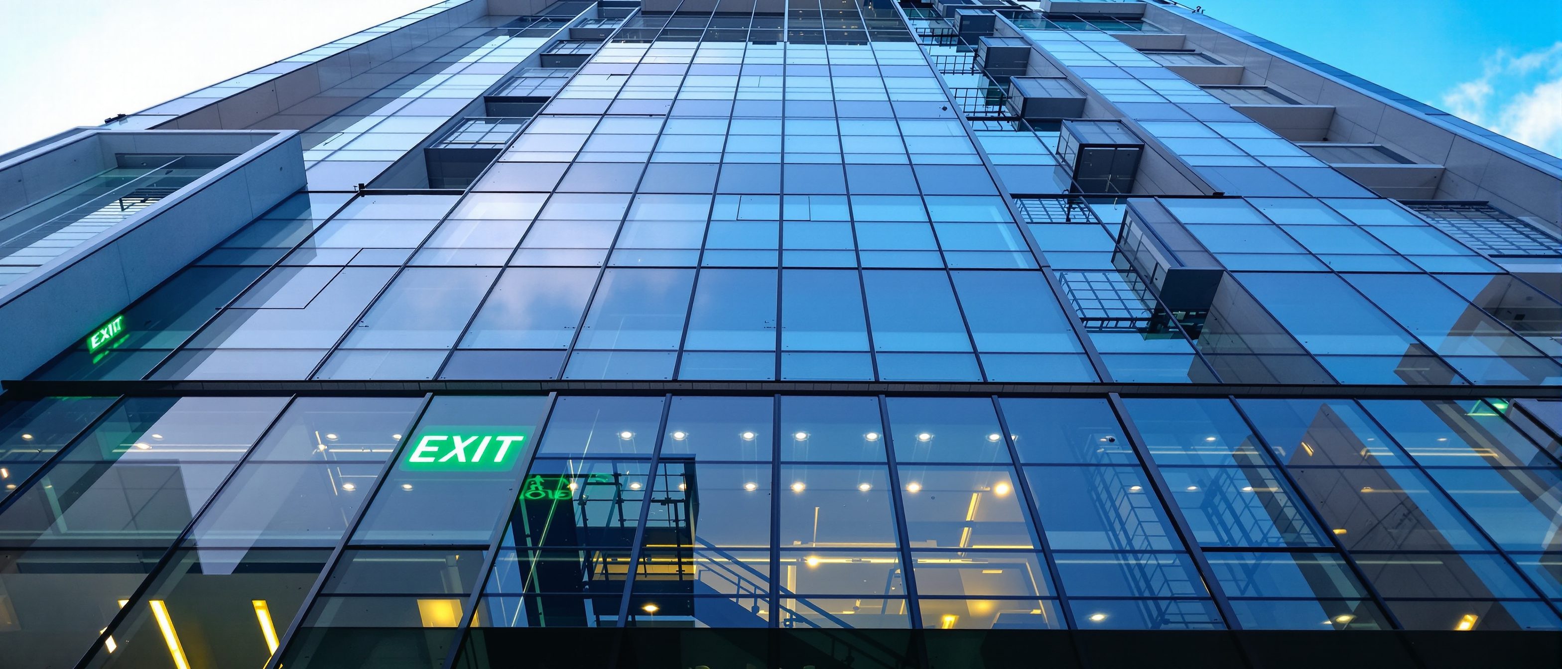 A modern Indian high-rise building seen from below against the sky, with an illuminated green emergency EXIT sign and a protected fire staircase visible, the life-safety provisions a tall building must have.