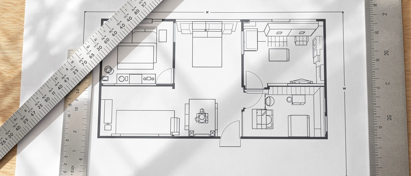 A dimensioned furniture-layout plan of a one-bedroom apartment on a drafting desk — walls, a sofa, bed, dining table and kitchen drawn to scale with a scale rule and set square, warm daylight, no people, no legible text.