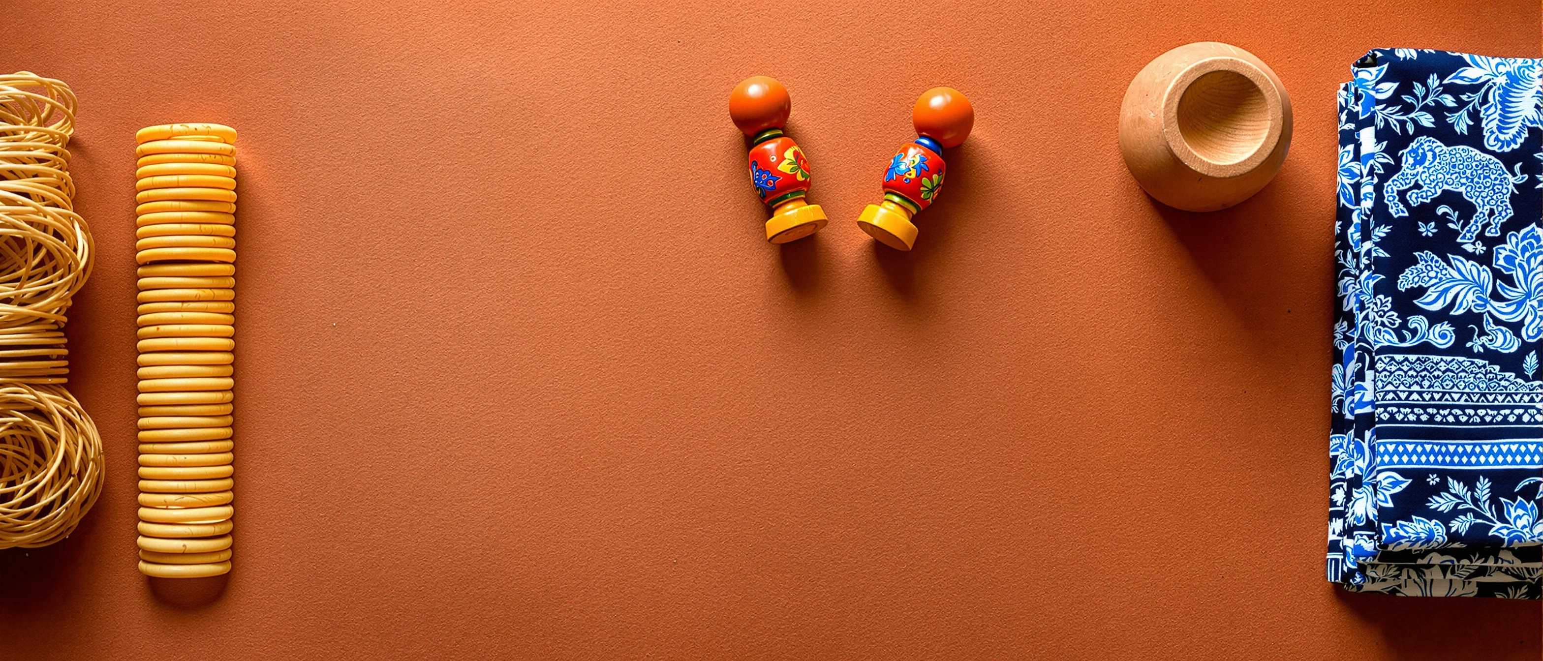 An overhead flat-lay of Indian craft objects — a block-printed indigo textile, a brass lost-wax figure, painted wooden toys and a terracotta horse — on a warm earthen surface