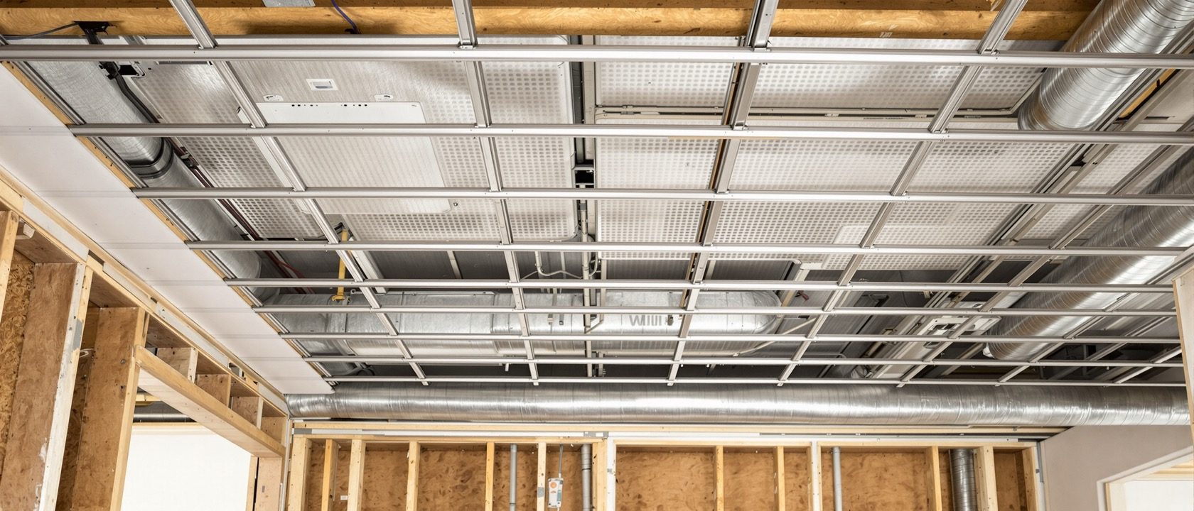 An interior ceiling under construction — a suspended GI metal framing grid with gypsum boards being screwed up on one side and an exposed 600x600 metal grid ceiling on the other, with AC ducts and wiring visible in the plenum above, warm site light, no people, no legible text.