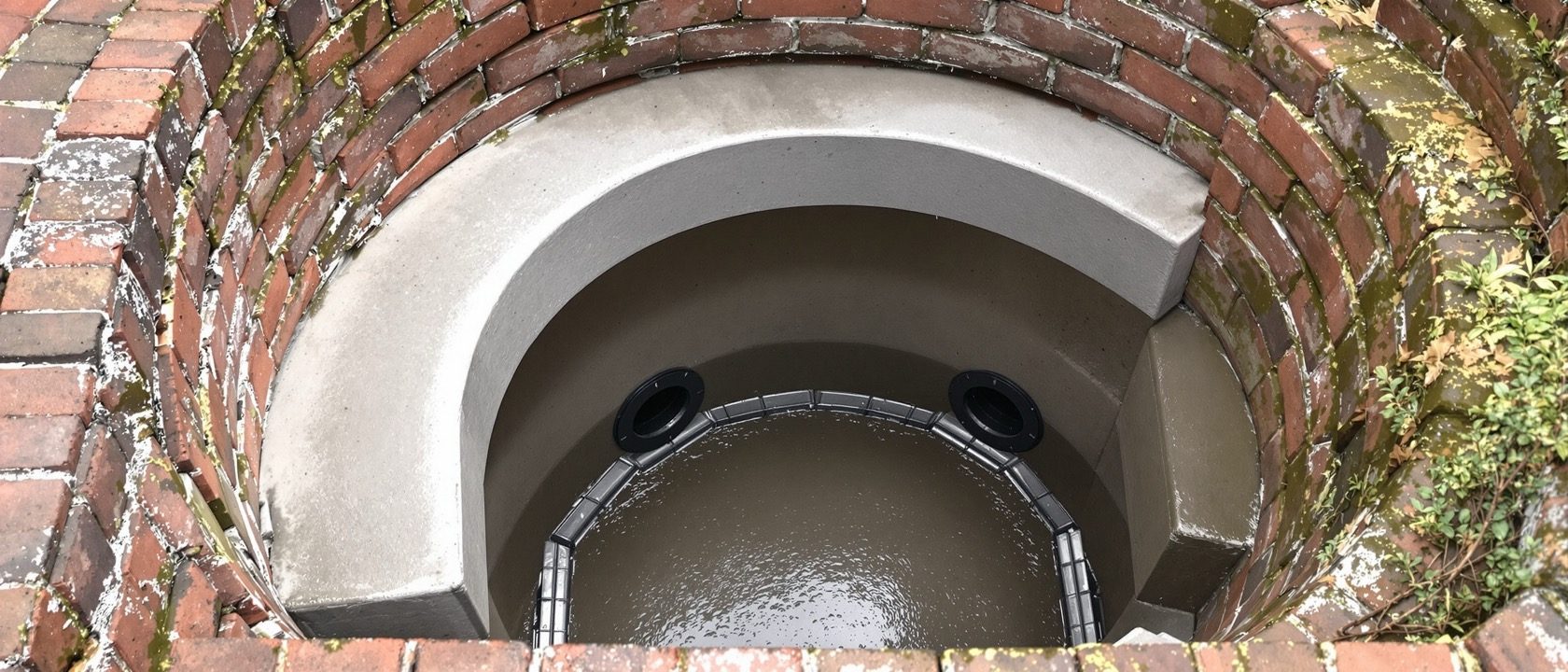 An open brick inspection chamber with a smooth benched channel and cast iron drain pipes entering it, seen from above in daylight