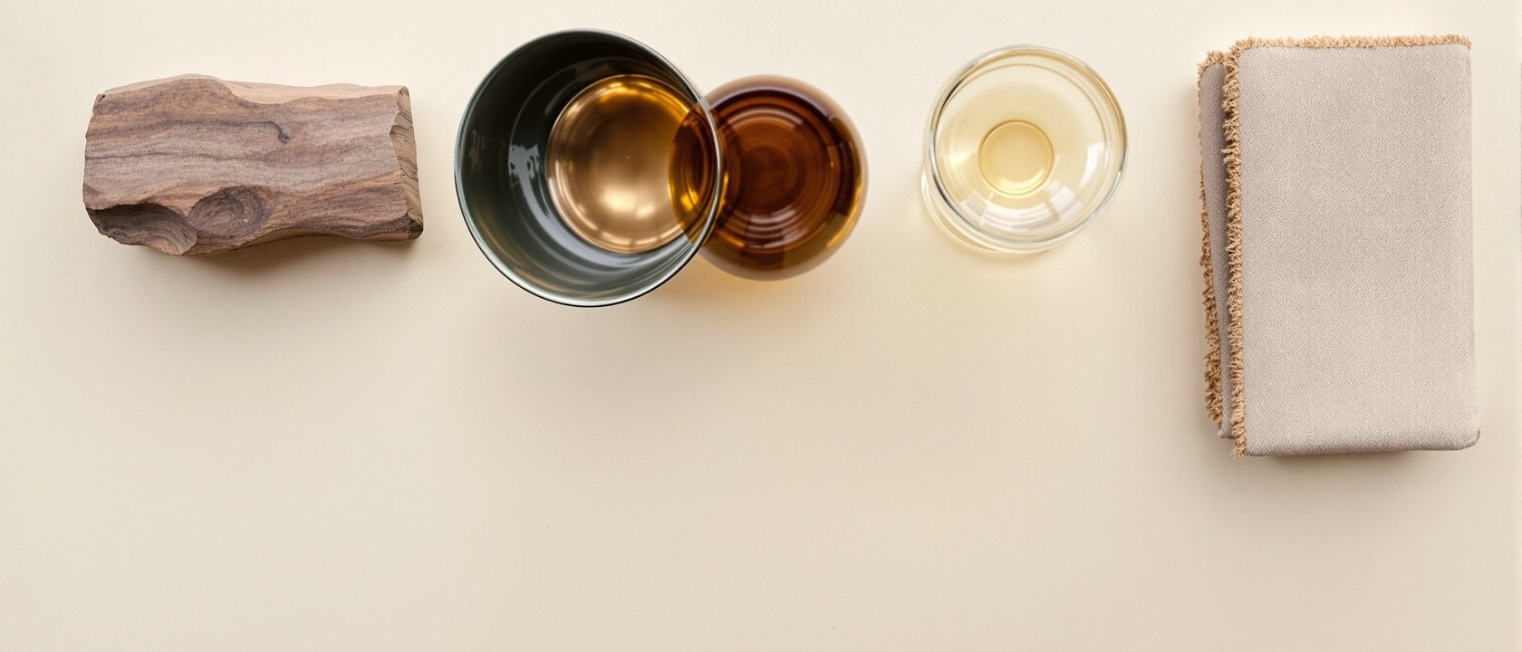 A flat-lay of accessory material samples on a plain surface — turned wood, brass, blown glass, glazed ceramic and folded handloom textile — tactile and warm, no text