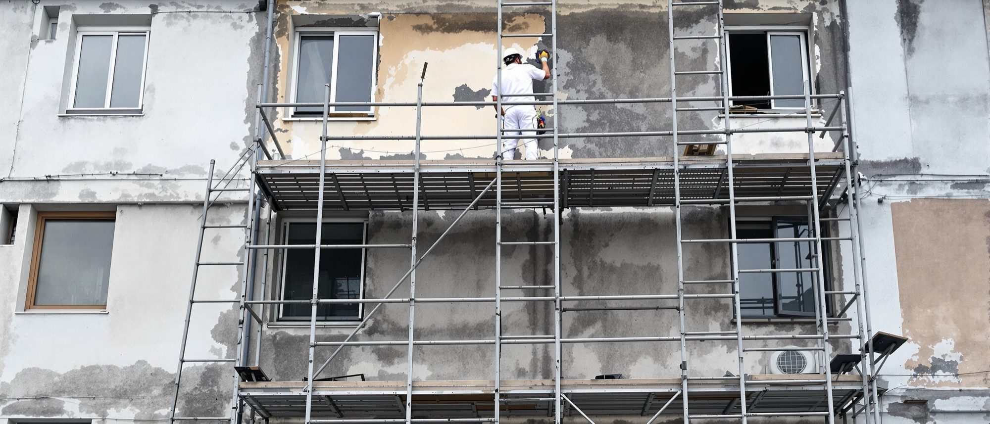 A building facade under maintenance — scaffolding against a weathered wall and a worker repairing it: keeping a building is the work of its whole life.