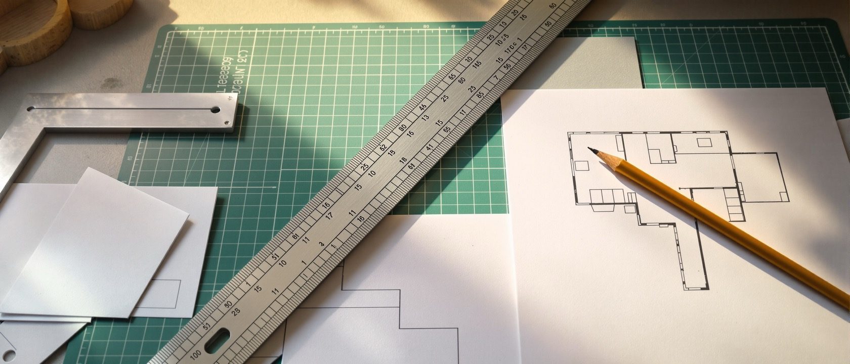 A model-maker's desk from above — a metal scale rule, a set square and a pencil laid over flat cut card templates of walls and a floor plan on a green cutting mat, warm daylight, no people, no legible text.