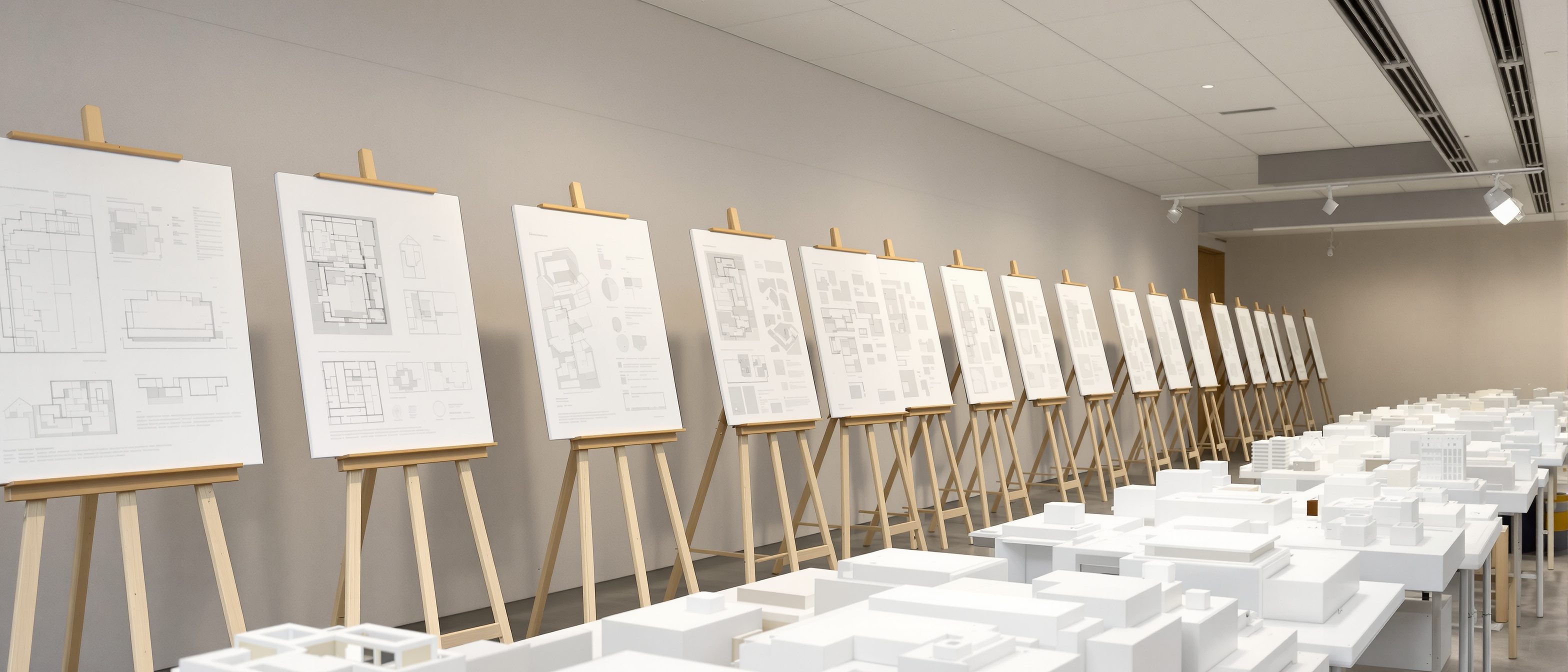 An architectural competition jury at work in India — anonymous entry boards with design drawings and models lined up along a wall, a panel of assessors studying them, the merit-based contest where the best design wins the commission.