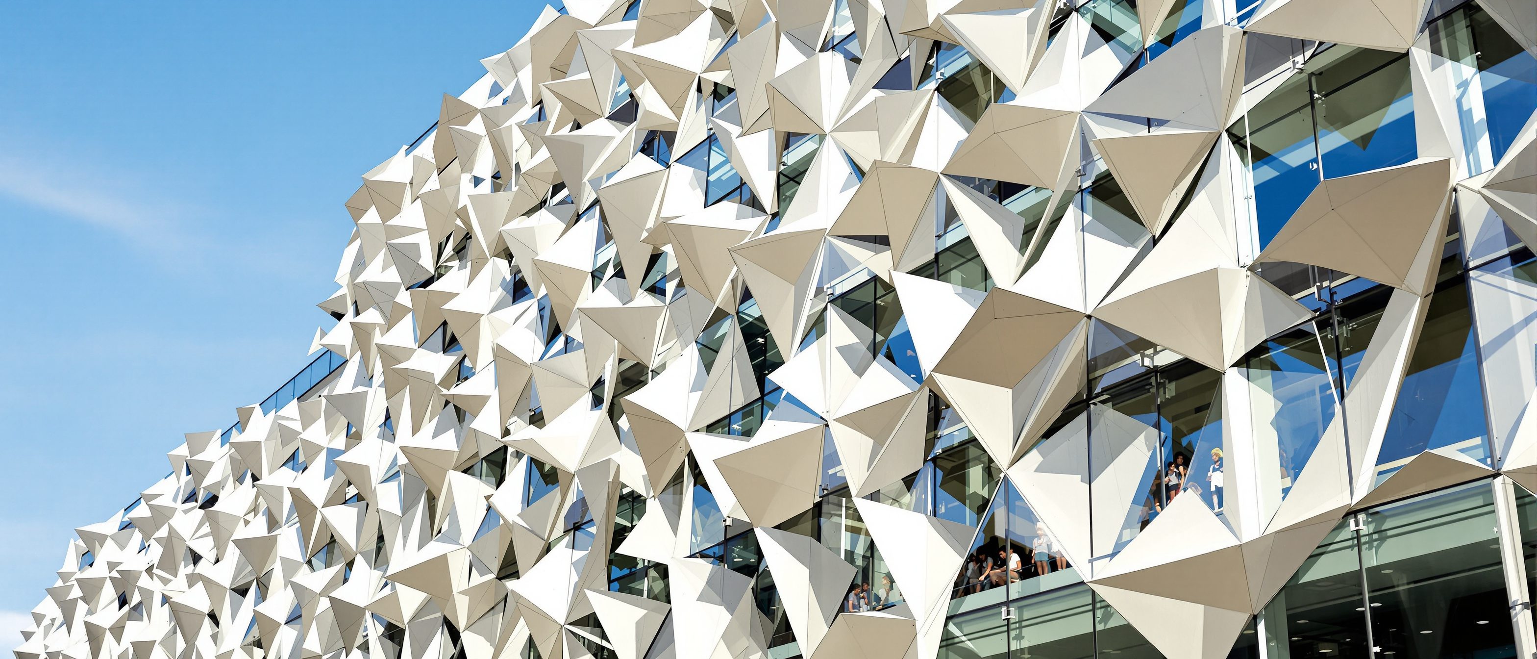 A responsive kinetic facade of triangular folding shading modules, some open and some closed across the glass — shade that tracks the sun, in the mashrabiya tradition revived as high-tech.