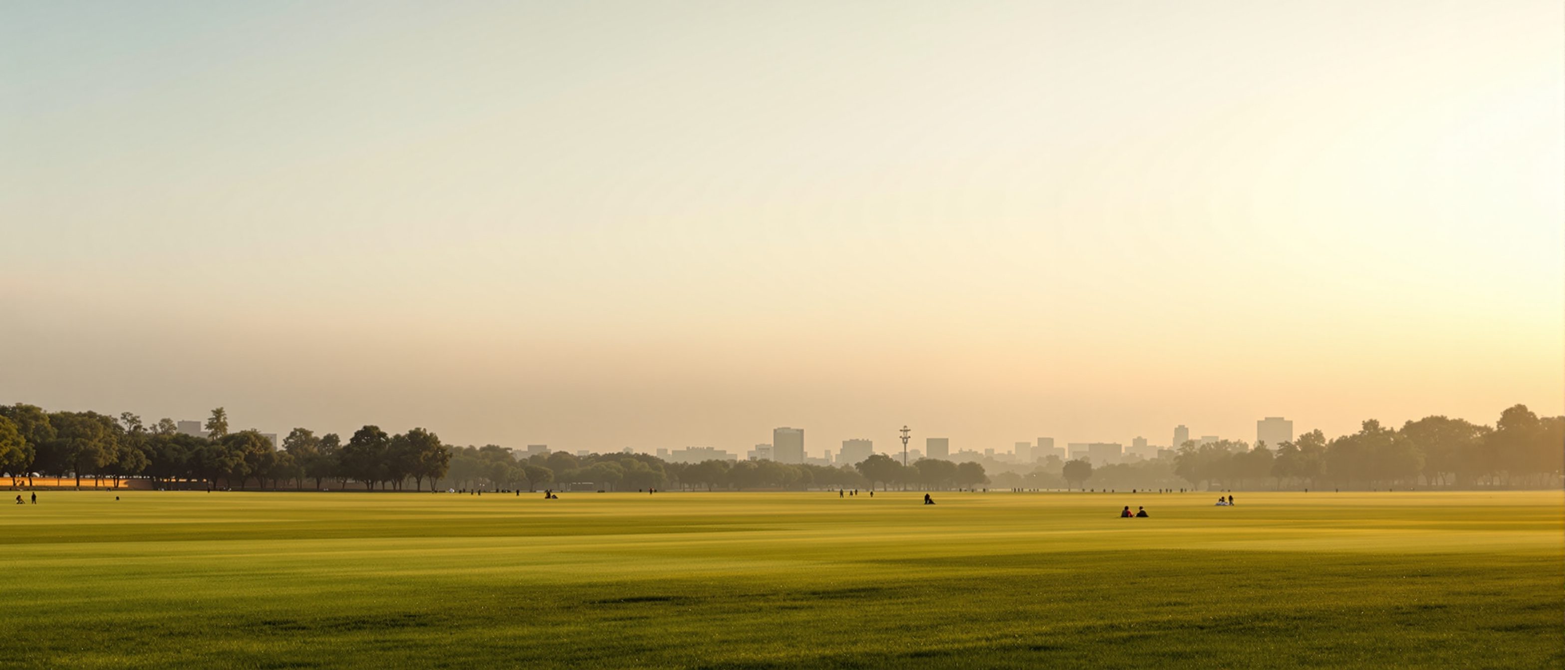 A large open Indian maidan at golden hour — a vast public green ground with people walking, sitting and playing, framed in the distance by the city skyline: the collective lung of the Indian city.