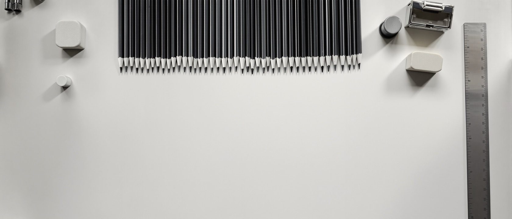 A drawing-studio desk with a graphite pencil-grade study — a stepped tonal ramp in pencil, a set of pencils from hard to soft, a sharpener and an eraser on white paper, warm daylight, no people, no legible text.