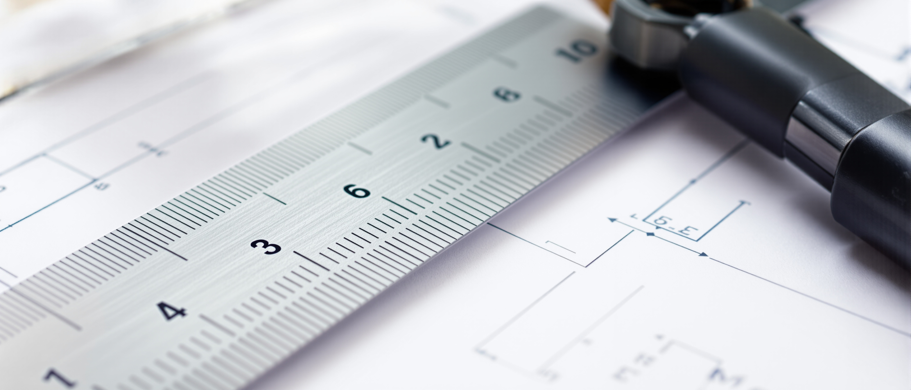 A triangular metal scale ruler and a clear set-square resting on a sheet of white drafting paper with faint plain pencil lines, close and technical in even daylight, no text